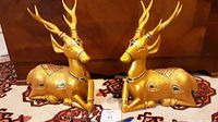 Side view of two large golden deer wood carvings with colored glass inlays and detailed antlers on wooden surface.