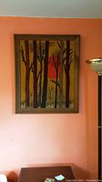 Framed textile crewelwork art depicting vertical tree trunks with an orange sun or moon in background, hung on orange wall above small table and floor lamp.