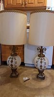 Pair of Hollywood Regency style lamps with white globular bases and cream fabric lampshades with gold rope trim, standing on kitchen floor in front of cabinets