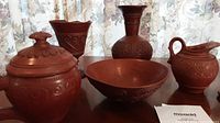Five old terracotta pottery pieces arranged on wood surface with a lace curtain background. Pieces include a lidded jar, tall vase, goblet, bowl, and pitcher.