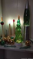 Photo showing silver glittery lava lamp, green glass Christmas tree lamp illuminated, bulb electric candles, and gold/green Christmas balls piled together