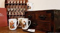 Photo showing six Franklin Mint collectible spoons on display rack, four Norman Rockwell mugs in front, and rustic wooden storage box beside them
