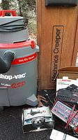 Side view of gray and red Shop Vac with 'Wet/Dry Vacuum' and 'QSP' branding, with mechanic's creeper and other tools around.