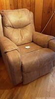 Front angled view of tan fabric upholstered recliner chair showing cushioned backrest, armrests, and seat with a folded note on the seat.