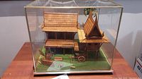 Full front view of the Thai house model inside glass case showing roof details, surrounding greenery, wooden structures, and base.