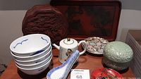 Overall view: carved red wood panel, lacquer tray, bowls, soup spoons, teapot, metal dish, celadon box and red lacquer dish