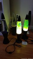 Five vintage lava lamps on wooden floor with power cords attached, lit showing various colors of lava in green, yellow, and clear liquid. One lamp is missing the cone top and one missing a bulb.