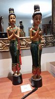 Two tall wooden Thai Sawasdee ladies praying figurines standing on a table in front of a decorative mirror.