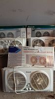 Three white window fans stacked against a wall, two Duracraft brand and one Cool Breeze Twin Window Fan