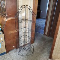 Full view of black metal corner wire shelf rack standing on carpeted floor near other wooden furniture and doorway.