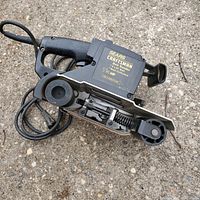 Photo of a black and gray Sears Craftsman electric belt sander laid on pavement surface, showing side profile with label clearly visible.