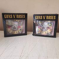 Two boxes of Guns N' Roses shot glasses side by side, each containing four shot glasses with band logos and themed designs. One unopened box and one used but new open box with slight damage.