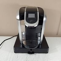 Front view of black and silver Keurig 2.0 coffee maker placed on top of black drip tray with drawer, showing digital display and silver accent details.