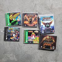 Photo shows six video games: Frogger (PS1), Rayman (PS1), Pac-Man World (PS1), Doom (computer), Doom Collector's Edition (computer), Ratchet & Clank Future (computer) arranged on concrete floor.