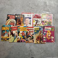 Collection of 11 vintage comics and magazines laid out on a concrete floor showing cover art and titles.