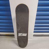 Top view of skateboard showing black grip tape with white Tony Hawk logo.