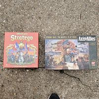 Photo showing box covers of Ultimate Stratego and Axis & Allies board games sitting on pavement.