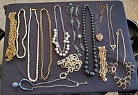 Twelve vintage-style costume jewelry necklaces laid flat on black fabric showing variety of styles, materials, and colors.