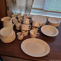 Full set of white ceramic dinette plates, cups, bowls, pitcher, sugar bowl, and creamer on a dark wood table.