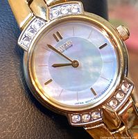 Close-up clear image of the watch face showing mother of pearl dial, gold-tone hands, hour markers, and crystal accents on the watch case.