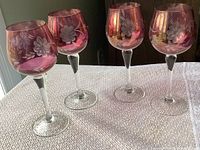 Four cranberry pink wine glasses with etched floral cornflower designs on bowls, clear stems and bases, placed on a textured light surface.