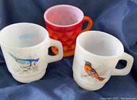 Three Fire King mugs displayed on dark blue fabric. Two stackable white mugs with bird designs on the front and one orange Kimberly Diamond textured mug behind.