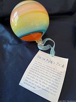 Glass Friendship Ball blending orange, blue, and yellow, featuring a tag explaining its meaning.