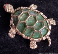 Close-up image of the turtle brooch showing the green stone shell with gold hexagonal trimmed segments, textured gold legs, head, and tail, and red rhinestone eyes.