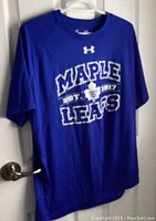 Full front view of blue Under Armour men's T-shirt with large 'MAPLE LEAFS' text, the established year 1917 and the Toronto Maple Leafs logo in white on the chest. The shirt is hanging on a hanger on a door knob.