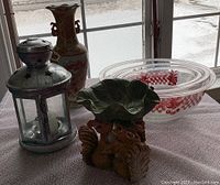 Photo showing items on a table near a window: metal lantern, decorative floral vase, ceramic squirrel dish with leaf bowl, and three clear Pyrex glass bowls with red cherry/checkered pattern.