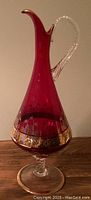 Full view of the tall red glass pitcher showing shape, gold detailed design and clear pedestal base.