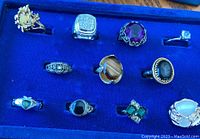 Blue cushioned display case holding 12 assorted rings, showing variety in style and color.