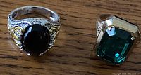 Two rings on wooden surface. Left ring silver-colored with black stone and gold-colored crab/lobster accent. Right ring gold-colored with large emerald green stone and intricate band.
