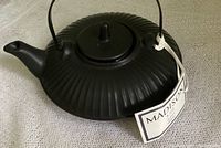 View of the black ribbed ceramic teapot with handle, spout, lid, and attached Madison & Co. branded tag showing the front and left side.