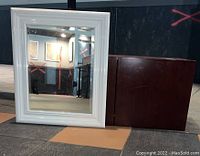 Photo showing the dark reddish-brown wood storage cabinet with double doors, positioned next to the white plastic framed mirror leaning on a wall.