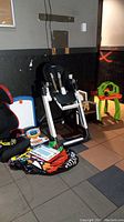 Photo showing Peg Perego highchair, Graco car seat, wooden rocking chair, green plastic easel/desk, and several toys and children accessories on floor