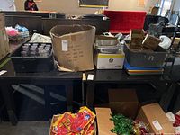 Wide view of various kitchen and serving supplies on black table including boxes, trays, plastic tub, and cutting boards.