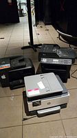 Three printers placed on tiled floor: HP OfficeJet Pro 8710, HP OfficeJet Pro 9015, Brother MFC printer. Visible condition used, cords partially seen.