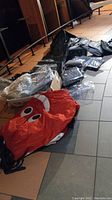 Overview of lot showing deflated red inflatable man with eyes/mouth, various black bags and pillow covers on tiled floor