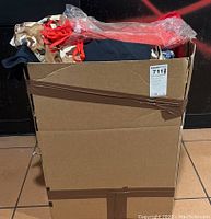 Cardboard box full of assorted clothing items including tops, corsets, hats, and other assorted apparel piled inside.
