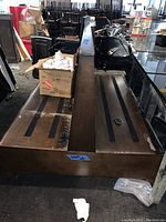 Photo of one wooden double-sided bench seat viewed from top and one end, showing seating surfaces with black strips and surrounding area with boxes and chairs.