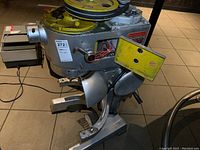 Side view of large gray commercial mixer with yellow pulley wheel on top and yellow cover plate detached and placed on side. Visible wiring and mechanical parts.