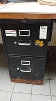 Front view of black metal two-drawer file cabinet, showing handles, label holders, wear and rust along bottom edge.