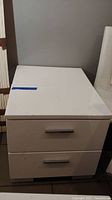 White laminate Coaster nightstand with two drawers, showing scratches, stains, and blue tape on top