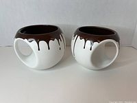 Two round white ceramic mugs with a brown drip design around the rim shown side by side on a white background.