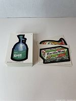 Photo showing two stacks of Wacky Packages trading cards; one stack is face-up showing 'Draft Keggs' parody, the other stack shows 'Hardly-Wrap' parody visible on top cards.