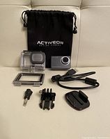 Photo showing Activeon Solar XG camera, waterproof case, rectangular frame mount, USB cable, two mounting brackets including adhesive mount, thumb screw, and black carrying pouch with Activeon branding.