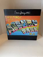 Photo of Bruce Springsteen's 'Greetings From Asbury Park, N.J.' vinyl album cover showing the colorful retro postcard-style design.
