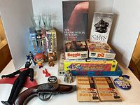Photo shows the entire lot including the books, board games, drinking glasses, figurines, replica pistol, and collectible pins and buttons arranged on a table.