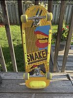 Full view of the vintage 1980s Snake skateboard standing upright outside on a wooden surface with green grass and a fence in the background. Shows deck, trucks, yellow wheels, and yellow rails.